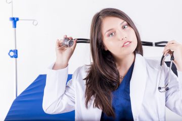 photo of a 30 something female doctor with long brown hair wearing a white lab coat and blue scrub top holding a stethoscope