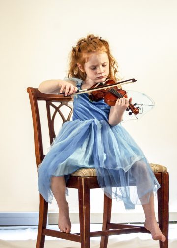 little red haired girl wearing a lacy blue dress sitting on a brown kitchen chair playing on a small violin