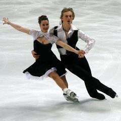 white couple in their twenties wearing matching black and white outfits figure skating arm in arm while they glide on ice