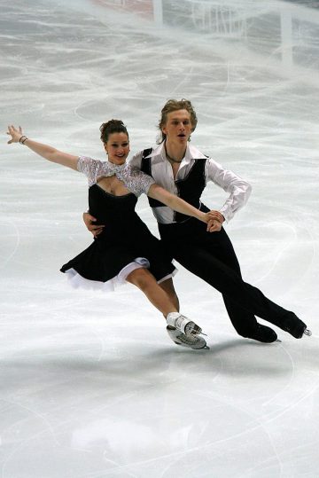 white couple in their twenties wearing matching black and white outfits figure skating arm in arm while they glide on ice