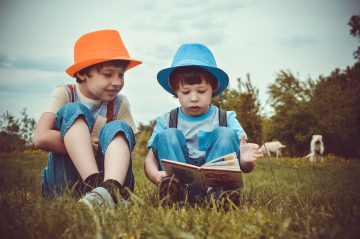 four year old boy with blue hat and six year old boy in orange hat reading a book together in a field with cows in the background