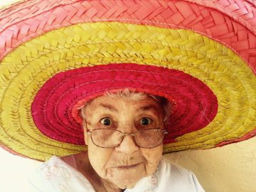 elderly lady with glasses wearing a huge red and yellow striped sombrero smiling into the camera