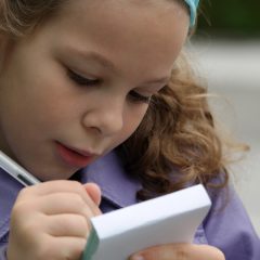 eight year old girl with sandy colored curly hair wearing a purple coat holding a memopad and pen