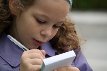 eight year old girl with sandy colored curly hair wearing a purple coat holding a memopad and pen