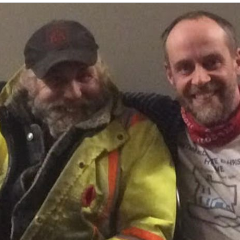 older man wearing a black ballcap and a yellow and orange work jacket sitting beside another middle aged man with a striped sweater and red bandana