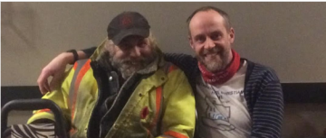 older man wearing a black ballcap and a yellow and orange work jacket sitting beside another middle aged man with a striped sweater and red bandana