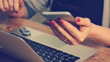 young woman hands with painted nails holding an iphone with her laptop in front of her