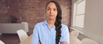 young woman in a blue blouse and thick brown hairbraid frowning
