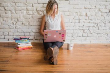 young blond woman sitting on the wood floor up against a white brick wall pink laptop on her legs