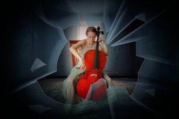 young woman in her twenties holding a big upright red cello and playing it with a bow while sitting on a chair